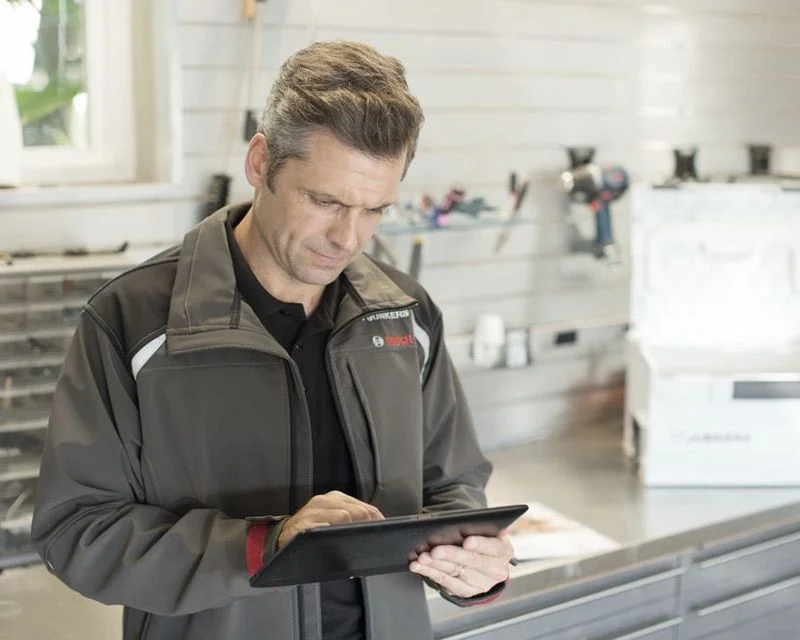 Ein Handwerker in Arbeitskleidung bedient ein Tablet in einer Werkstatt.